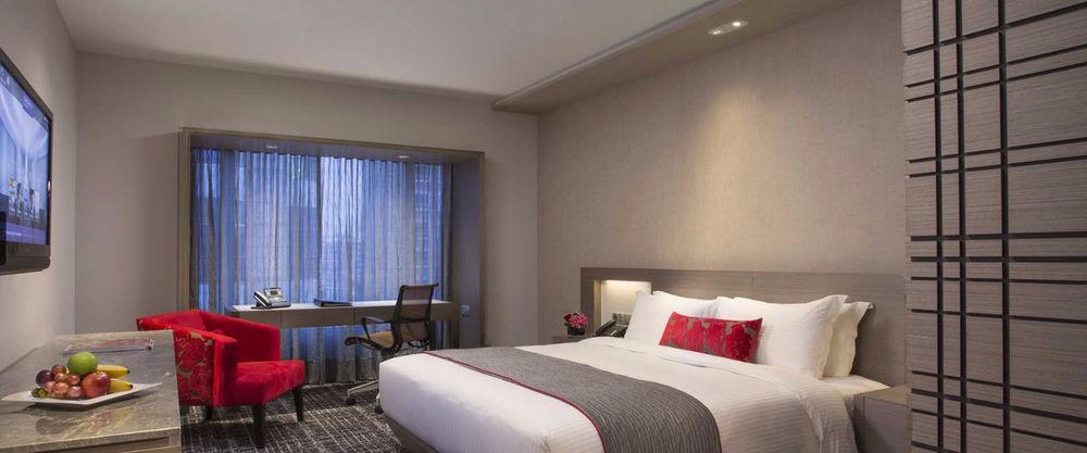 Carlton Hotel Singapore Esterno foto The photo shows a modern hotel room featuring a large bed with white linens and a gray bed runner. There is a decorative red pillow on the bed. To one side, a desk with a rolling chair is positioned near a window dressed with sheer curtains, allowing