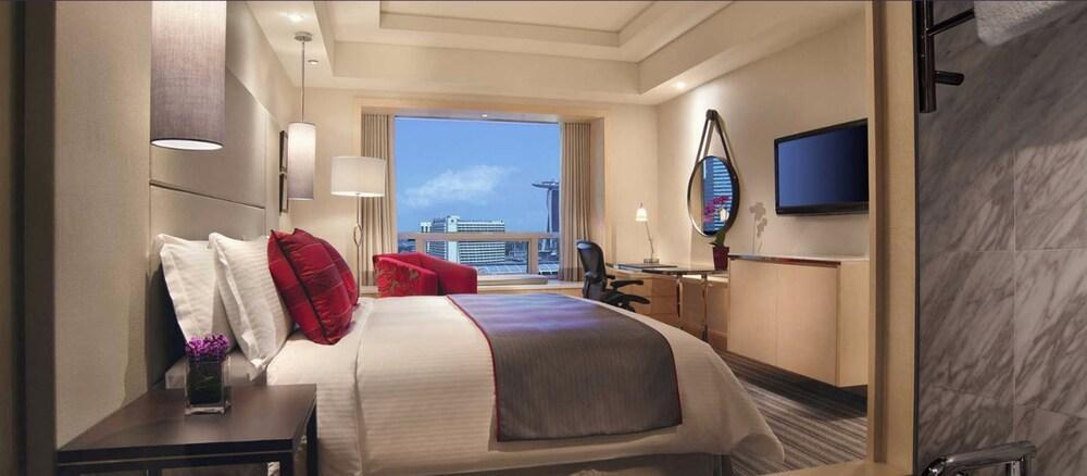 Carlton Hotel Singapore Esterno foto The photo shows a well-furnished hotel room. It features a large bed with white bedding and red accent pillows. There is a large window that provides a view of the outside, likely showcasing a cityscape or skyline. On the opposite wall, there is a mo