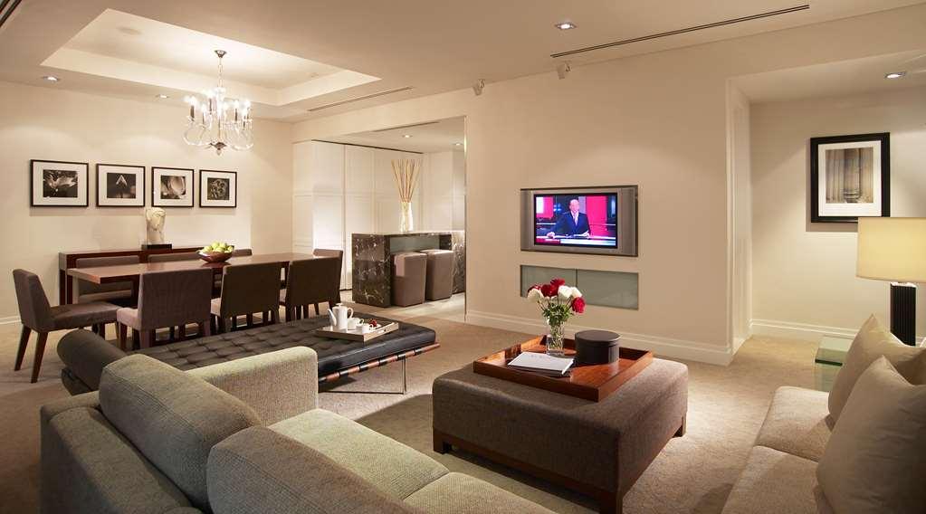 Carlton Hotel Singapore Camera foto The photo shows a modern living room that is part of an elegantly designed space. There are two gray sofas arranged in front of a coffee table, which holds a small vase of flowers and other items. A television mounted on the wall displays a news prog