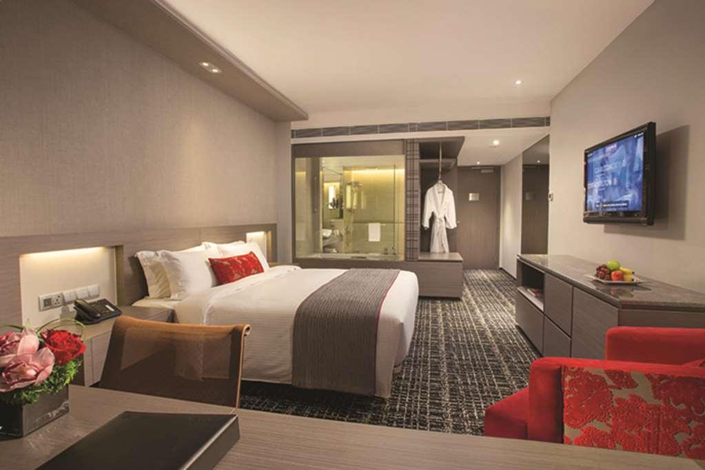 Carlton Hotel Singapore Camera foto The photo shows a modern hotel room featuring a neatly made bed with white bedding and red accent pillows. There's a contemporary desk with a black notepad in the foreground and a vibrant red chair beside it. A flat-screen television is mounted on th