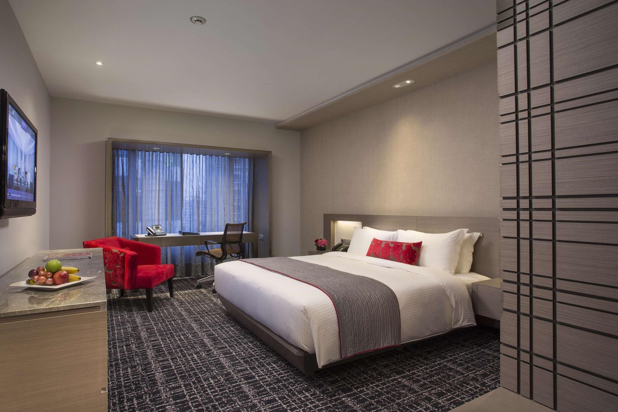 Carlton Hotel Singapore Esterno foto The photo shows a modern hotel room featuring a large bed with light-colored linens and a gray bedspread. There is a bedside table with decorative items and a small fruit bowl. On one side of the room, a red accent chair and a small desk are position