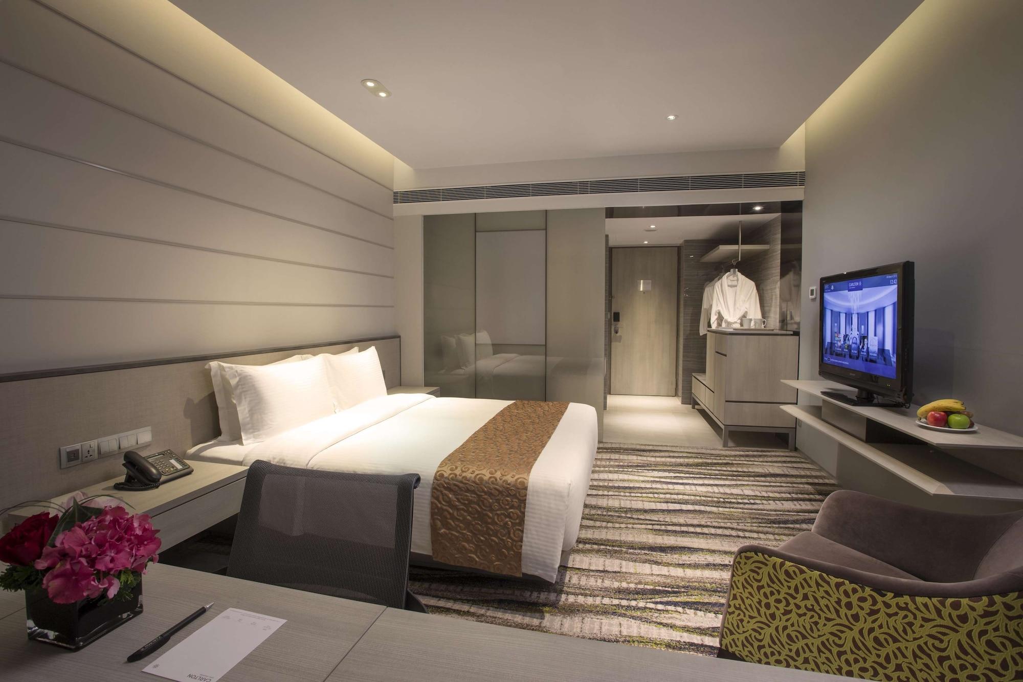 Carlton Hotel Singapore Esterno foto The photo shows a modern hotel room with a clean and contemporary design. It features a large double bed with white bedding and a decorative brown throw. There is a wall-mounted television across from the bed, and a small seating area with a chair. A