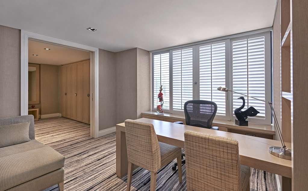 Carlton Hotel Singapore Camera foto The photo shows a modern, well-lit office space. There is a large desk with a sleek black office chair and two light-colored chairs on either side. The desk is situated near a large window with horizontal blinds, allowing natural light to filter in. 