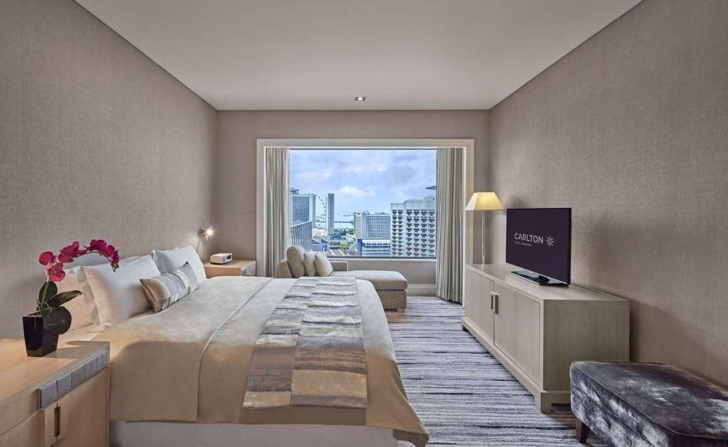 Carlton Hotel Singapore Camera foto The photo shows a modern hotel room featuring a large bed with a stylish headboard and neutral-toned bedding. There is a bedside table with a lamp and a small vase of flowers next to the bed. A sleek TV is mounted on the wall, and a comfortable seati