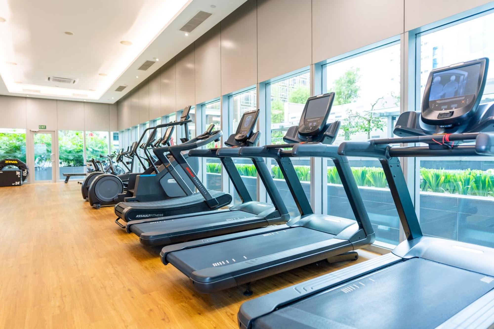 Carlton Hotel Singapore Esterno foto The photo shows a modern gym facility featuring several treadmill machines lined up in a row. The treadmills are positioned on a wooden floor and have digital display screens. Large windows provide natural light and a view of greenery outside, creati