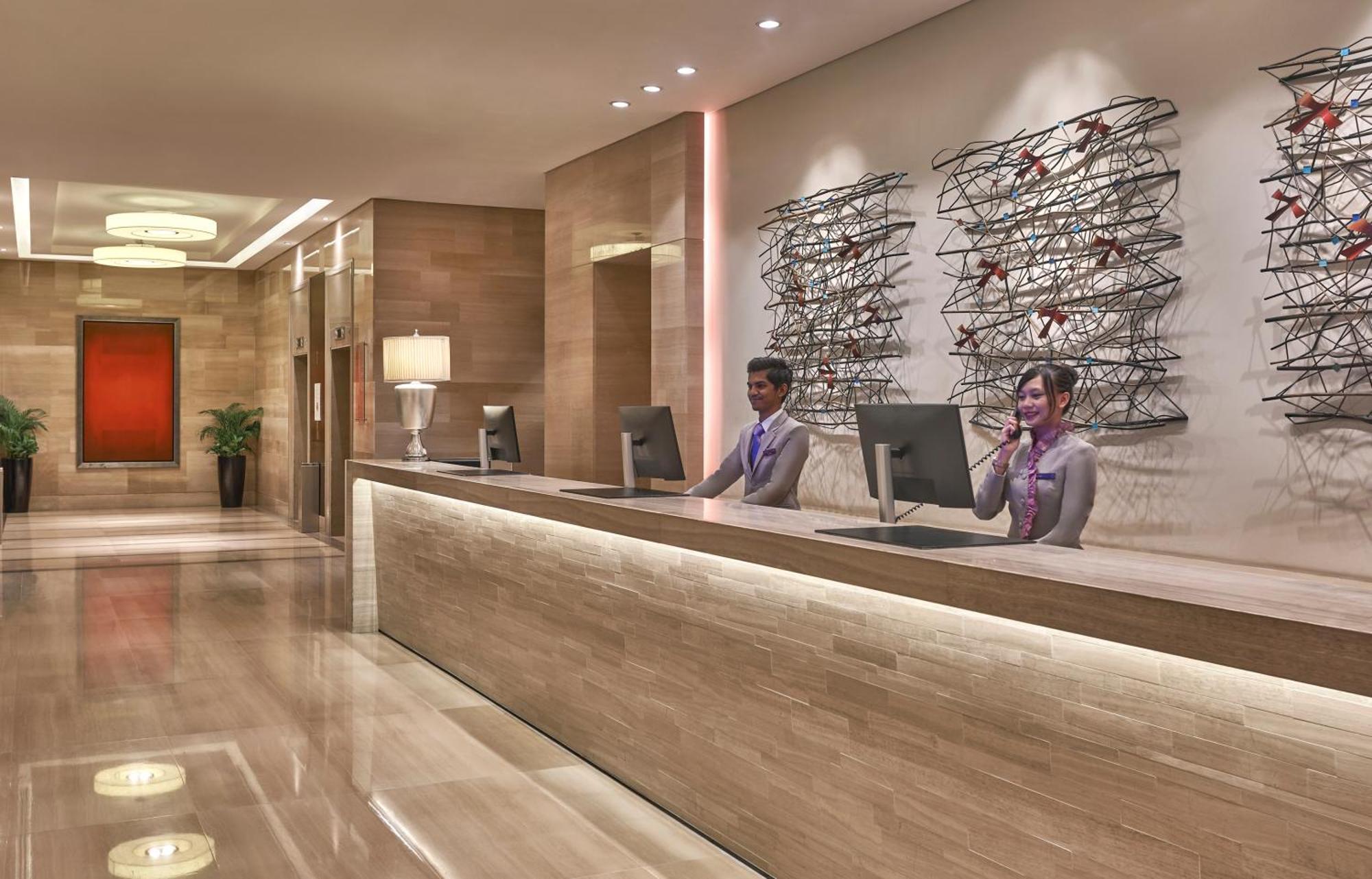 Carlton Hotel Singapore Esterno foto The photo shows a modern hotel reception area. In the foreground, there is a sleek reception desk made of light-colored stone, with soft lighting illuminating the front. Behind the desk, there are two receptionists engaged with guests, dressed in pro