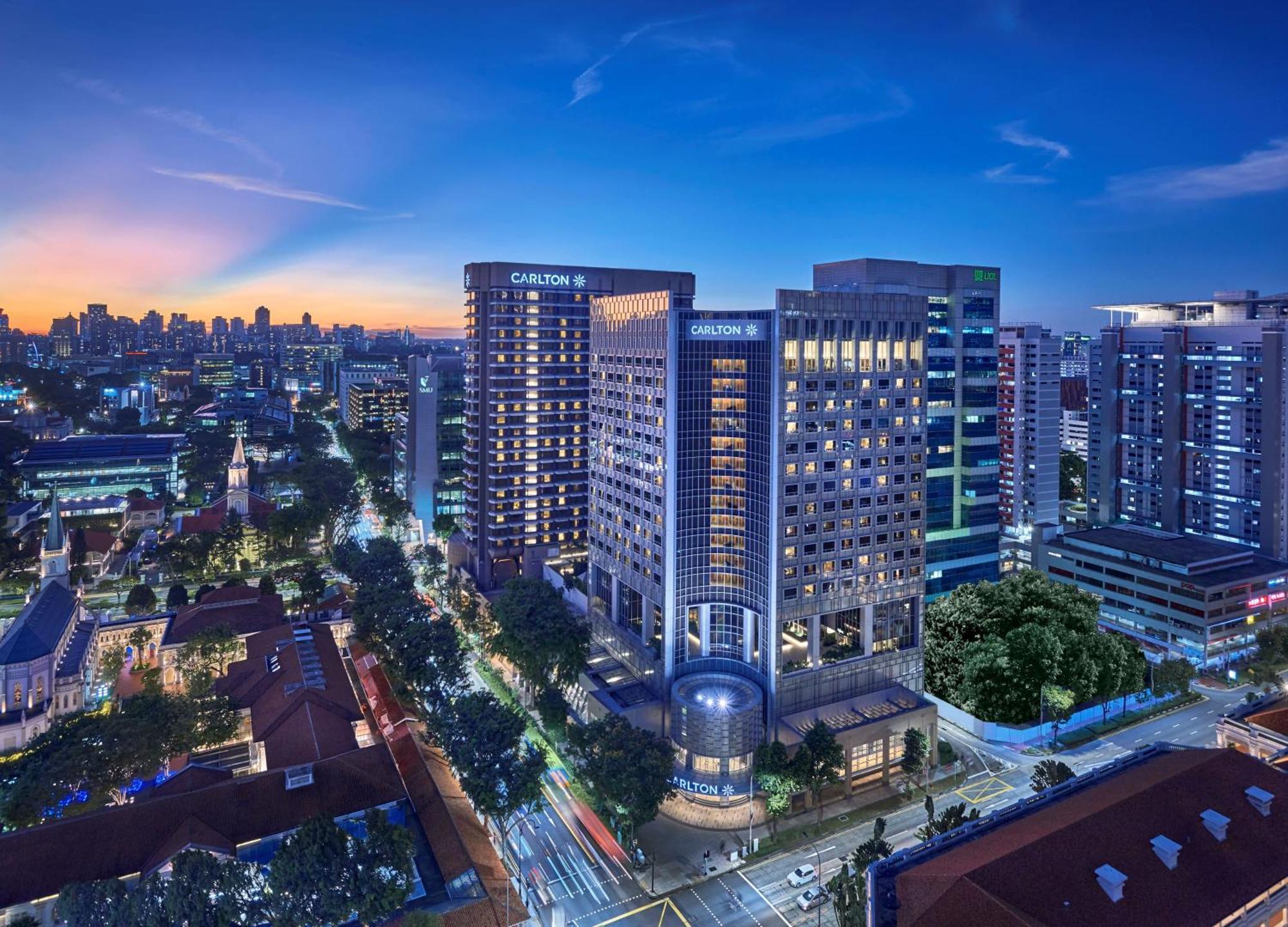 Carlton Hotel Singapore Esterno foto The photo shows a city skyline at dusk, featuring a cluster of modern buildings. Prominent in the image are several high-rise structures, including a large hotel that bears the name "Carlton." The scene is illuminated by city lights, and the sky tran