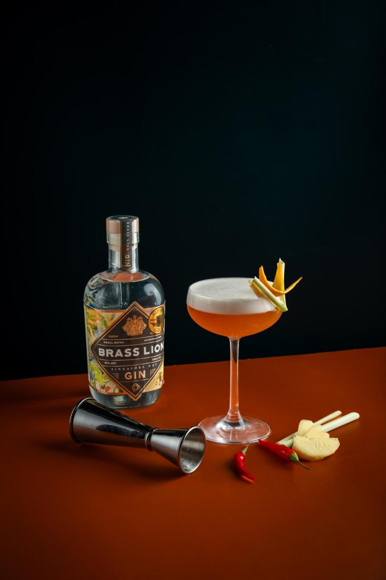 Carlton Hotel Singapore Esterno foto The photo features a bottle of Brass Lion gin prominently displayed alongside a cocktail. The cocktail is served in a coupe glass and appears to have a frothy top, likely from egg white or cream. It is garnished with colorful citrus peels and a slice