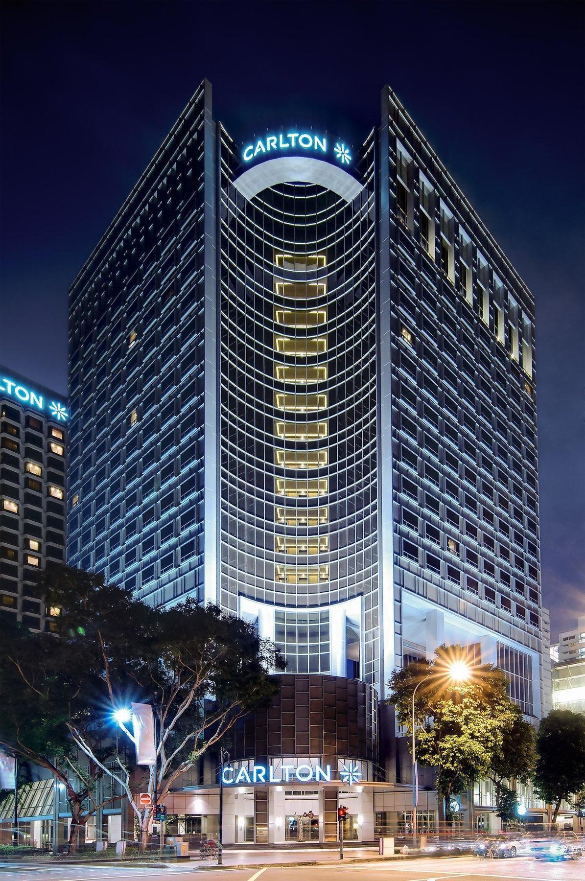 Carlton Hotel Singapore Esterno foto The photo shows a tall, modern high-rise building characterized by a sleek glass façade. The building has a distinctive curved design and features multiple floors with large windows, emitting a soft glow from the interior lights. It appears to be par