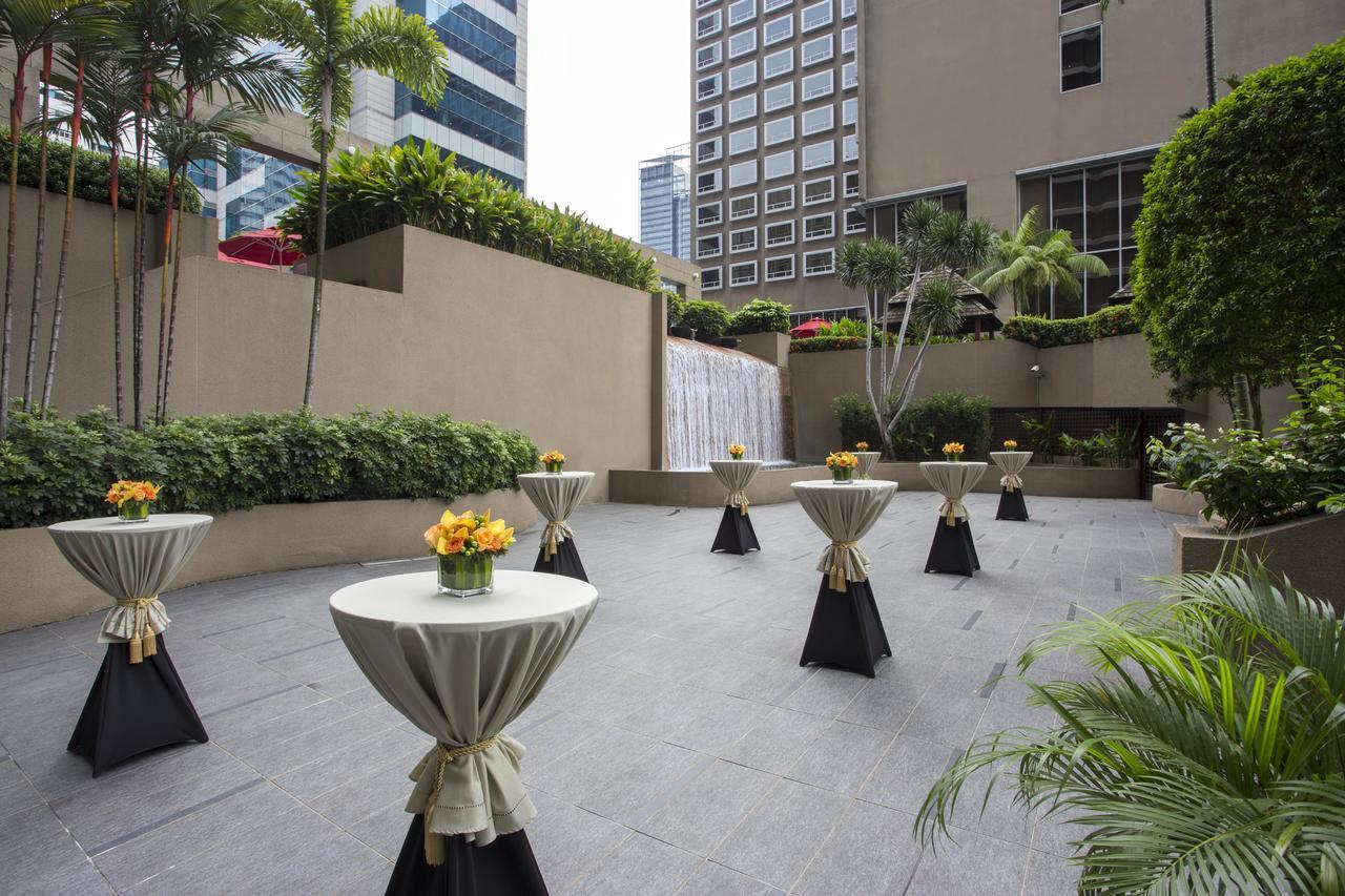 Carlton Hotel Singapore Esterno foto The photo shows an outdoor area designed for events or gatherings. There are several circular tables set up, each draped with a cloth and topped with a small arrangement of flowers in a vase. The backdrop features modern buildings, greenery, and a sm