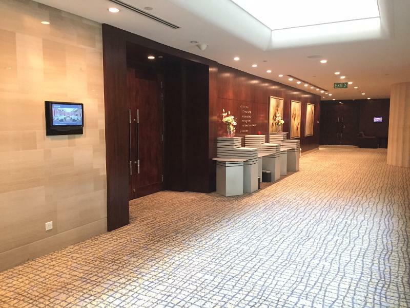 Carlton Hotel Singapore Esterno foto The image shows a corridor in a modern interior space. The hallway features a tiled floor with a patterned design, leading towards a well-lit area. On one side, there are several reception or information desks, which are paired with decorative flower
