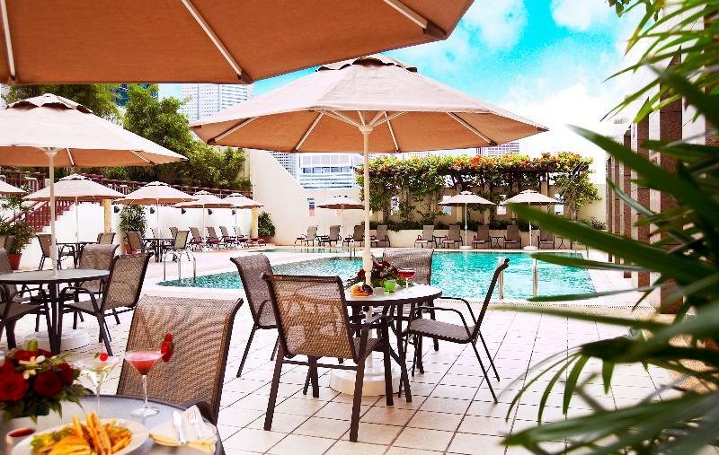 Carlton Hotel Singapore Servizi foto The photo shows a tranquil outdoor setting featuring a swimming pool surrounded by lounge chairs and umbrellas. There are tables with chairs on a tiled patio area, and some have drinks placed on them, suggesting a relaxing atmosphere. Lush greenery c