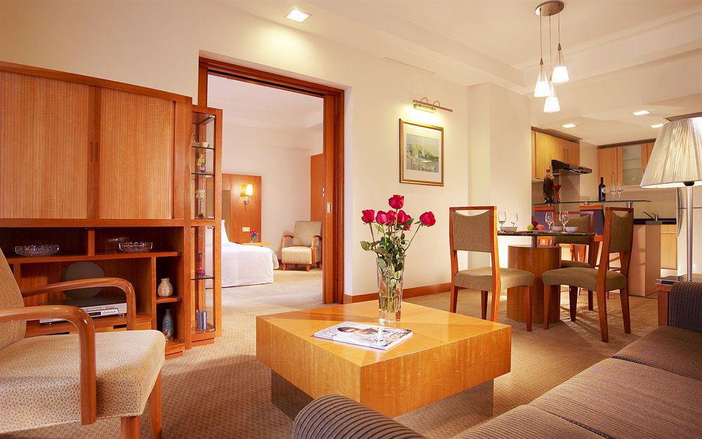 Carlton Hotel Singapore Camera foto The photo shows a cozy living space or suite. In the foreground, there is a small wooden coffee table with a magazine on it, surrounded by comfortable seating, including a sofa and armchairs. A vase of red roses adds a touch of color to the setting. 